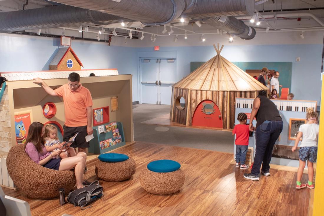 A group of adults and children engage with an exhibit, some of them sitting and reading, and others playing with a wall of musical instruments.