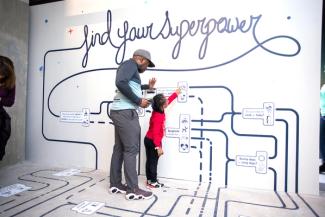 A maze winds onto a wall from the ground and has text saying "find your superpower." A man and child stand with their hands on the wall.