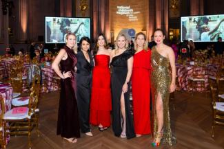 People in formal wear posing at the National Children's Museum Gala with tables, a stage, and large screens behind them