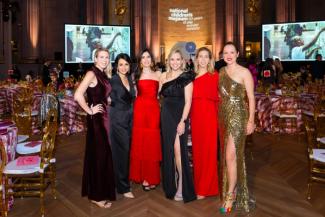 People in formal wear posing at the National Children's Museum Gala with tables, a stage, and large screens behind them