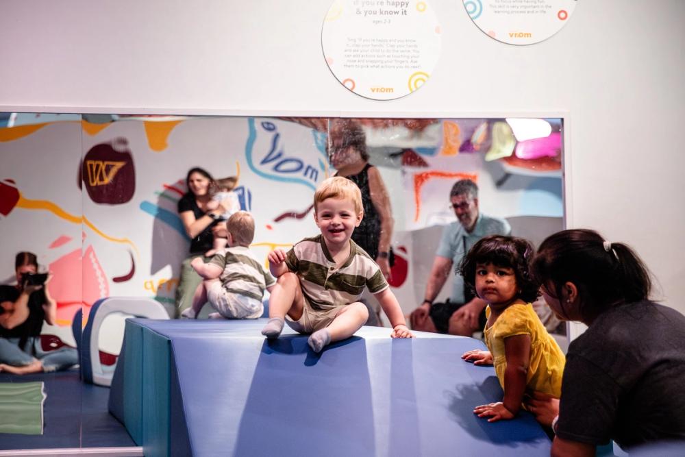 Two young children sit on foam gym blocks stacked in front of a large mirror.