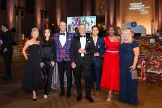 People in formal wear posing at the National Children's Museum gala with tables, a stage, and large screens behind them