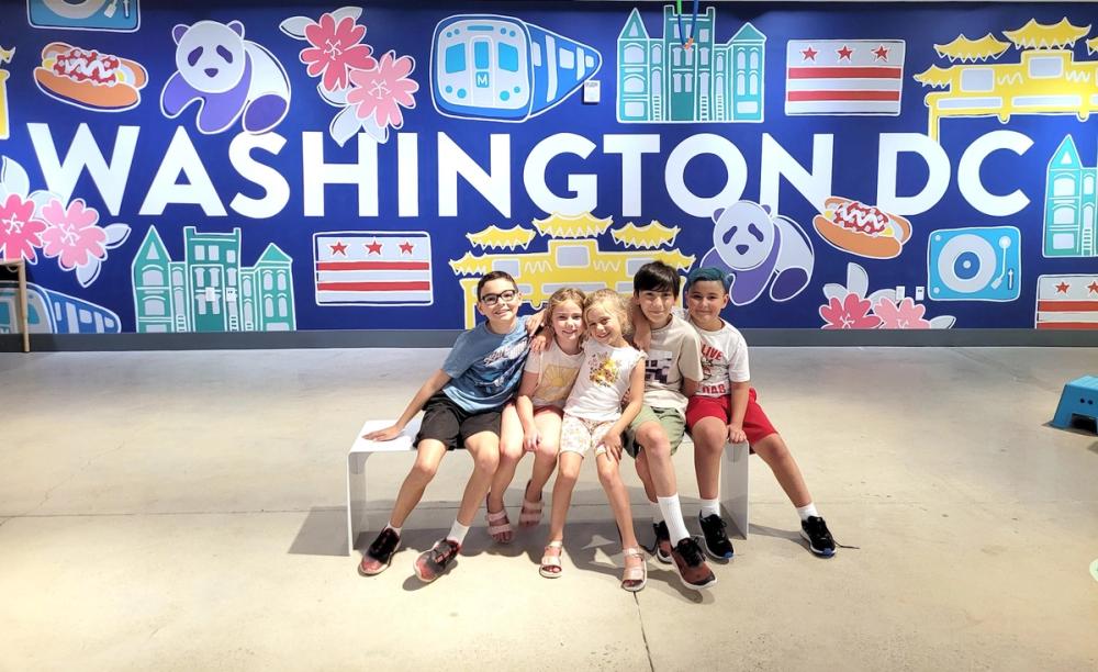 Five smiling children sit, squished, onto a white bench in front of a colorful mural that says "Washington DC."