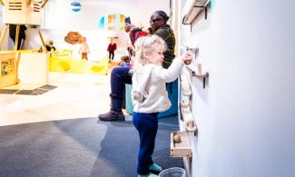 A toddler with blond hair places a wooden ball onto a wooden ramp mounted to a white wall.