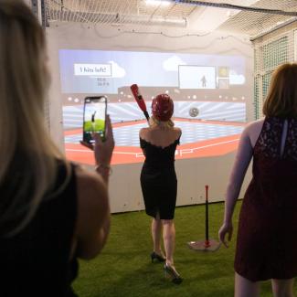 A woman in a formal dress plays digital baseball, wearing a helmet and holding a plastic bat, while other adults watch.