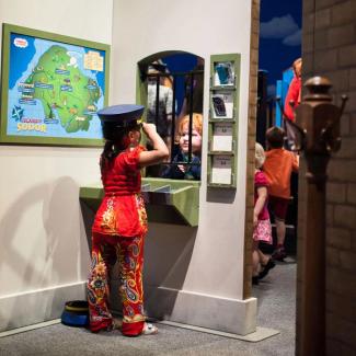 A little girl in a conductor hat at the ticket window with a little boy on the other side