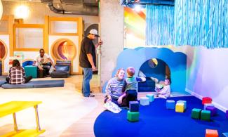 Toddlers play with colorful foam blocks on a play mat while adults sit on a play structure in the background.