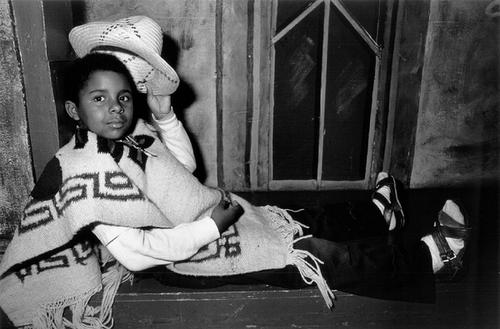 A young child reclines with legs extended, holding a straw hat in one hand, which is raised slightly off their head.
