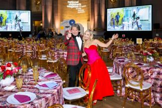 Two people in formal wear posing at the National Children's Museum Gala with tables, a stage, and large screens in the background.