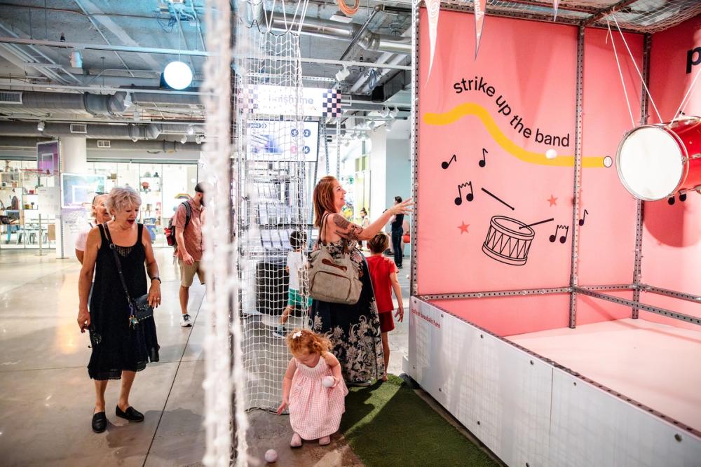 A woman throws a ball at a drum handing in a pink exhibit as a small child picks up baseballs.