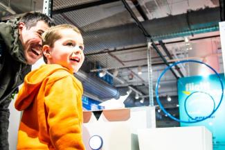 A toddler in an orange sweatshirt smiles with an adult man as they stand in front of a net with blue hoops hanging from it.