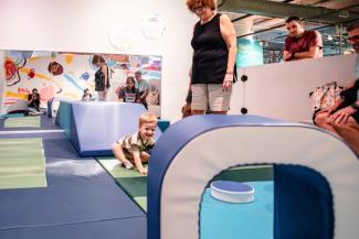 A young boy climbs on blue gym mats as adults watch, smiling.