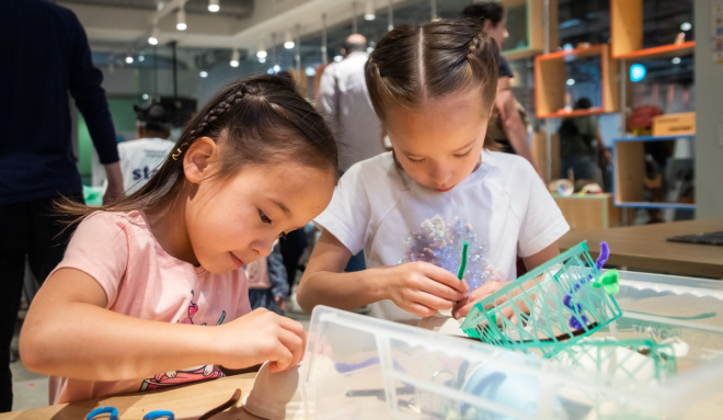 Two young girls are engaged in a craft activity at a table, focusing intently on their tasks. One girl is handling a crafting material, while the other works with a small green basket.