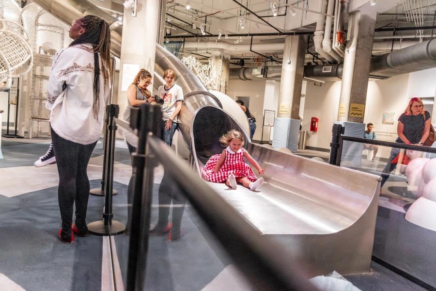 A young child in a gingham dress slides down a large metal slide past two adults.