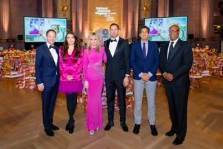 People in formal wear posing at the National Children's Museum Gala with tables, a stage, and large screens in the background.