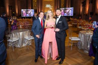 People in formal wear posing at the National Children's Museum gala with tables, a stage, and screens in the background