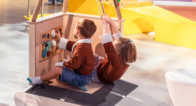 Two young children are playing at an indoor play area. They are seated in a wooden play structure with interactive knobs, gears, and ropes. One child reaches for a dial while the other pulls on a rope.