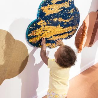 A small toddler in a yellow shirt reaches up to play with a beaded gold and blue cloud-shaped wallhanging.