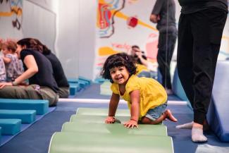 A toddler in a yellow shirt smiles at the camera as she climbs over a green wavy play mat.