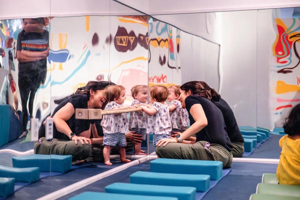 A woman sits on a gym mat watching her baby hold onto a wooden pull-up bar that is attached to a large mirror in front of them.