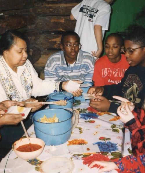 A group of people share a meal around a table in a rustic setting. An older woman on the left serves food from a blue pot.