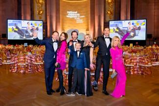 People in formal wear posing at the National Children's Museum Gala with tables, a stage, and large screens in the background.