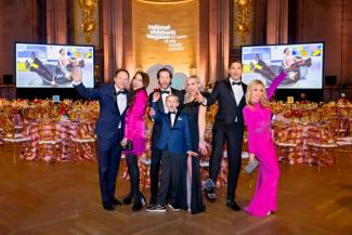 People in formal wear posing at the National Children's Museum Gala with tables, a stage, and large screens in the background.