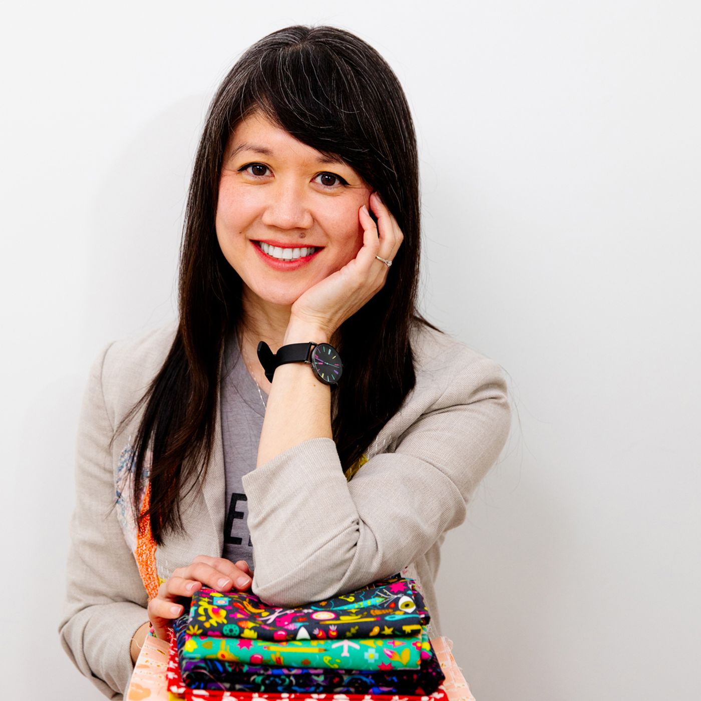 Diana Li faces towards the camera, with her chin in her left hand. Her left elbow is on top of a stack of brightly colored fabrics. 