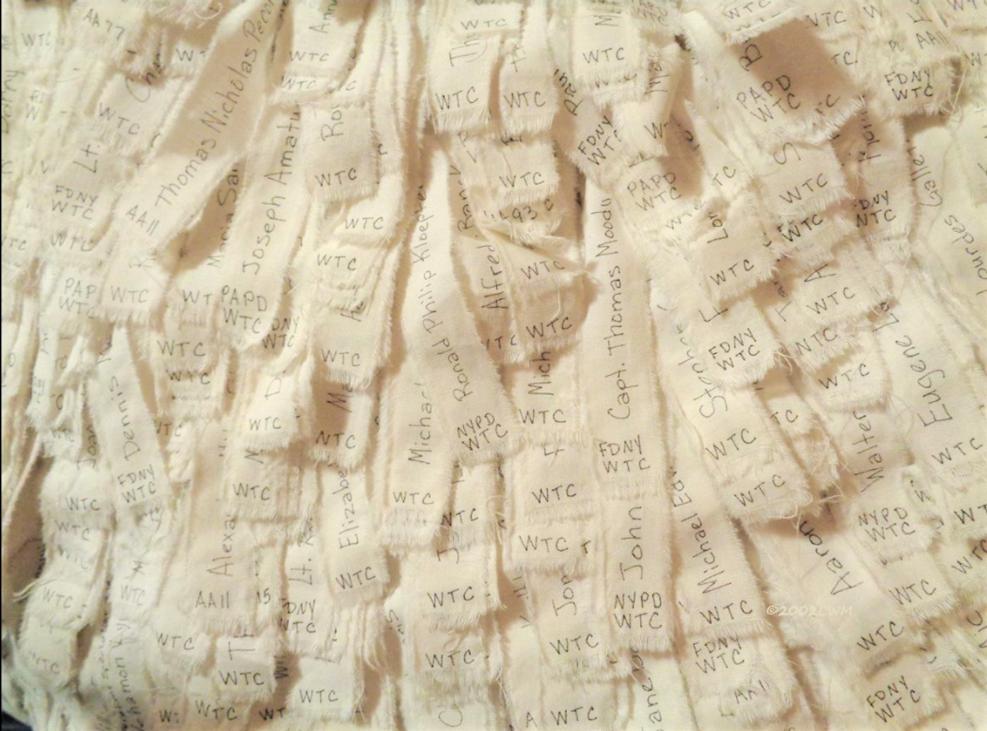 Hand-lettered fabric strips bearing the names of individuals lost on 9/11, each marked with a location such as WTC, FDNY, NYPD, or flight number.