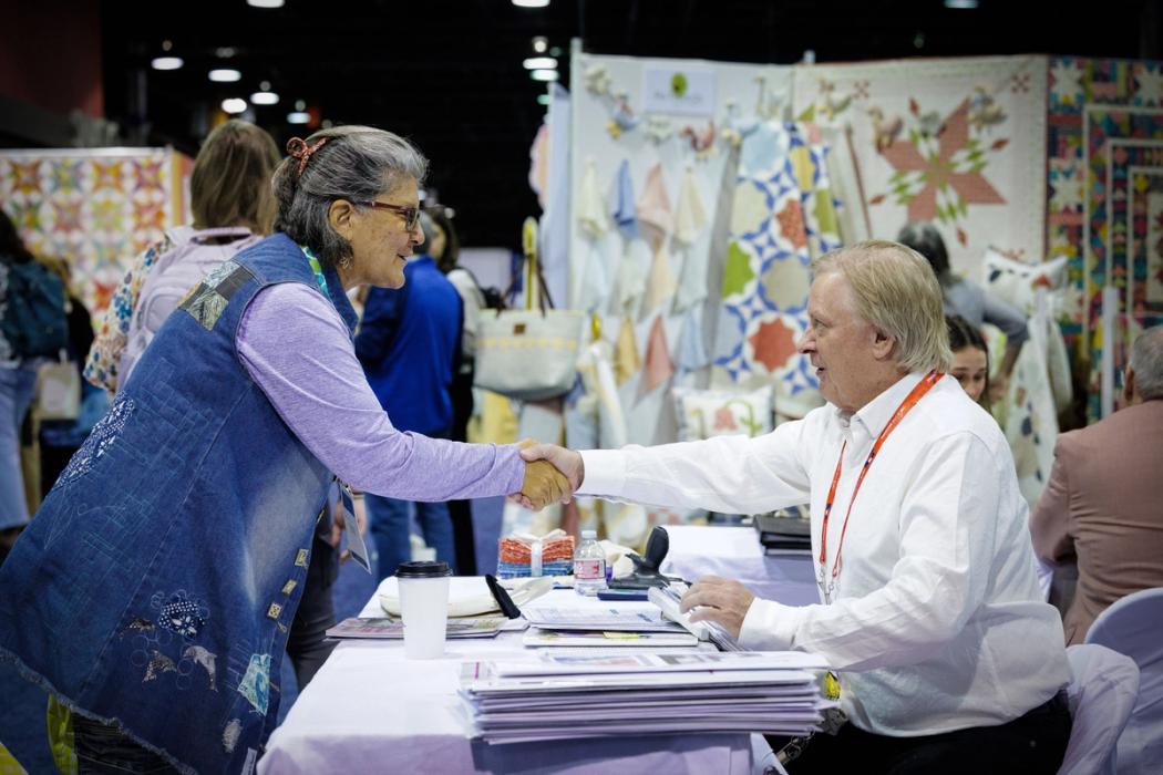 A standing woman shakes hands with a seated man on the trade show floor. 