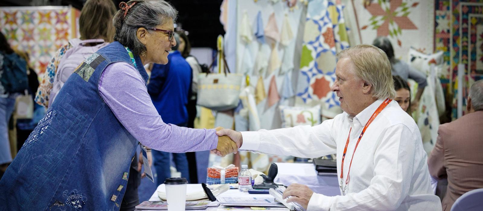 A standing woman shakes hands with a seated man on the trade show floor.