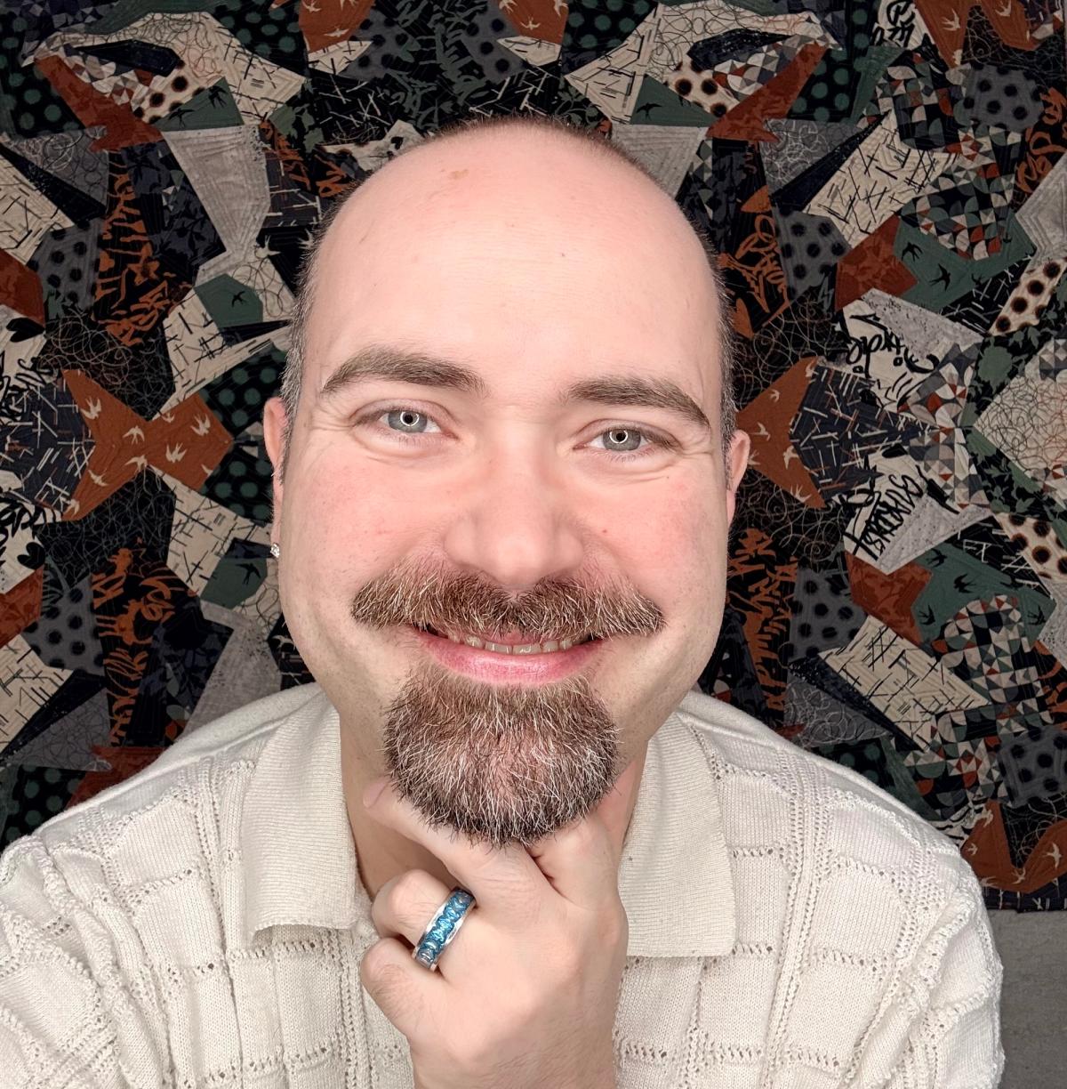 A man with a beard and mustache smiles at the camera, wearing a cream-colored sweater and a blue ring. A patterned quilt serves as the background. smiles at the camera, wearing a cream-colored sweater and a blue ring. A patterned quilt serves as the background.