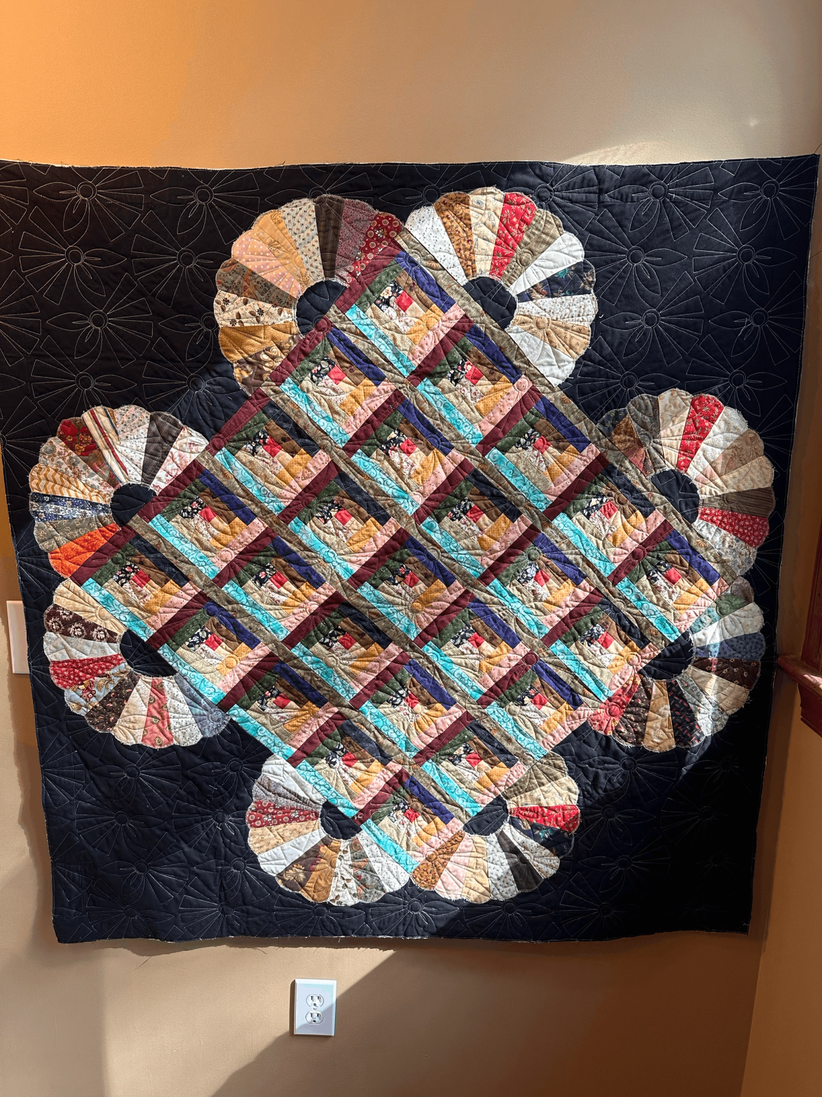 A large quilt featuring colorful diamond patterns in the center, surrounded by fan-shaped segments on a dark background, hanging on a beige wall above an electrical outlet.