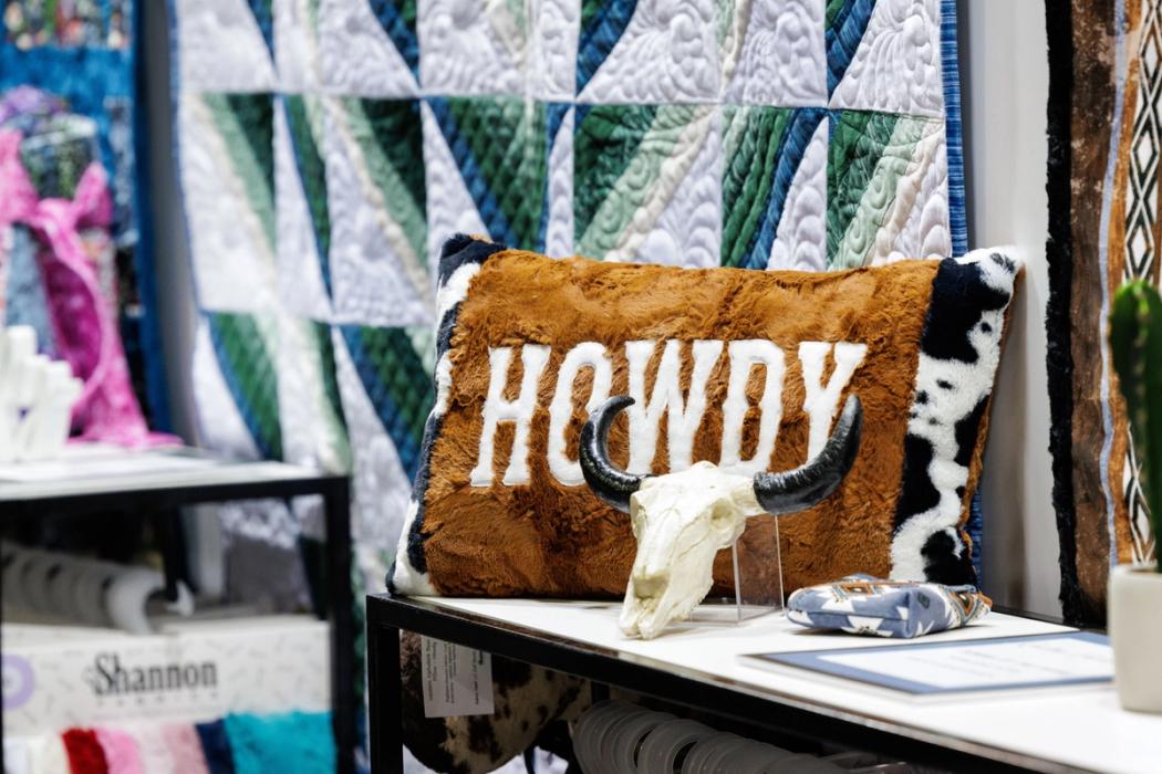 A brown pillow with the word "HOWDY" sits on a shelf next to a decorative cow skull, with colorful quilted fabrics in the background.