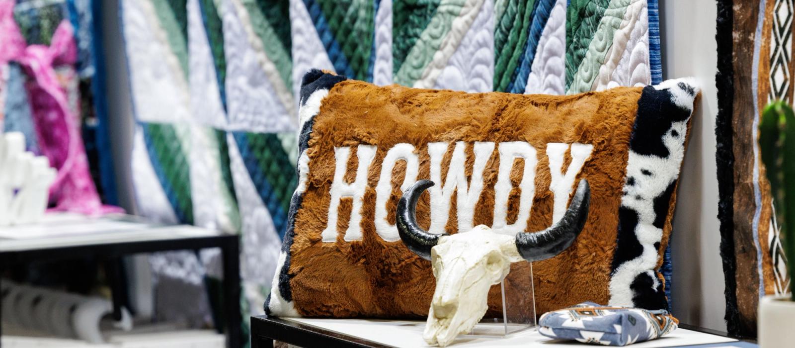 A brown pillow with the word "HOWDY" sits on a shelf next to a decorative cow skull, with colorful quilted fabrics in the background.