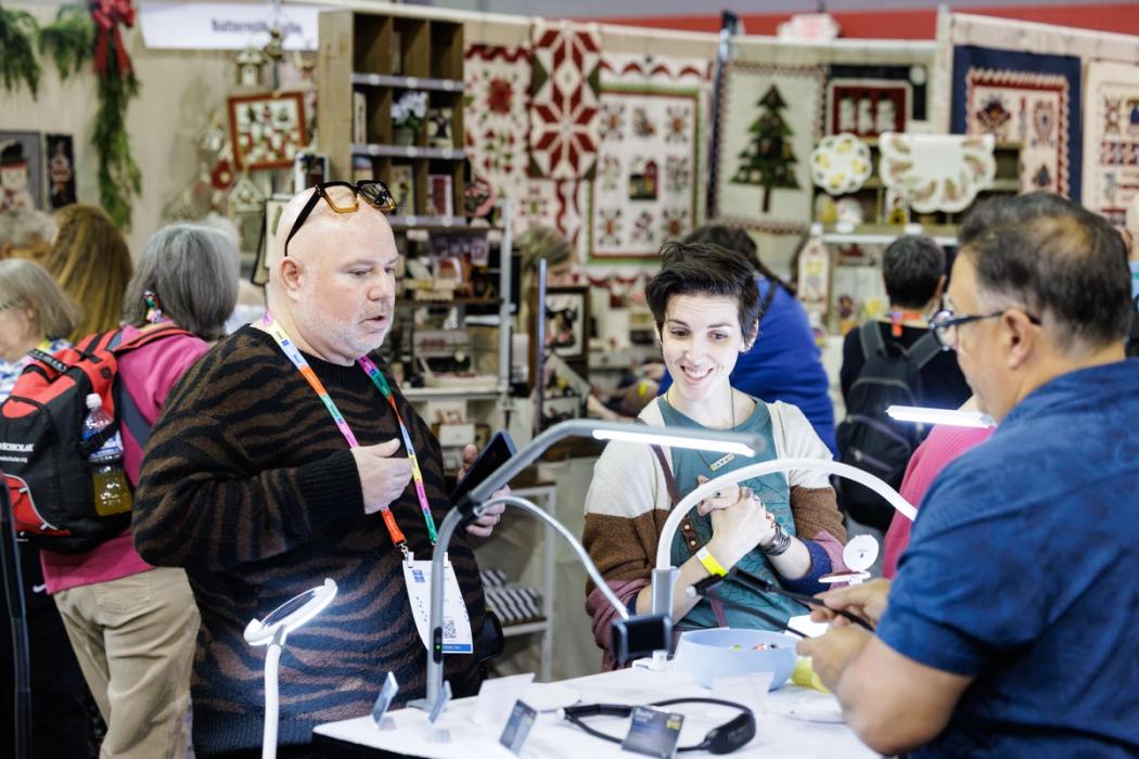 Three people interact at a craft fair booth featuring quilts, with a man in a tiger-print sweater talking to a woman and a man behind the counter.