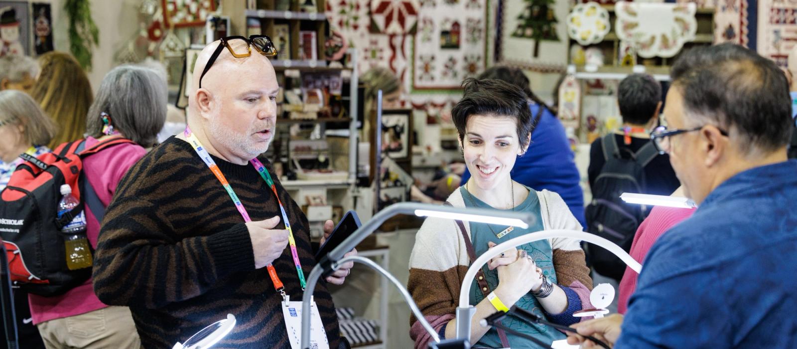 Three people interact at a craft fair booth featuring quilts, with a man in a tiger-print sweater talking to a woman and a man behind the counter.