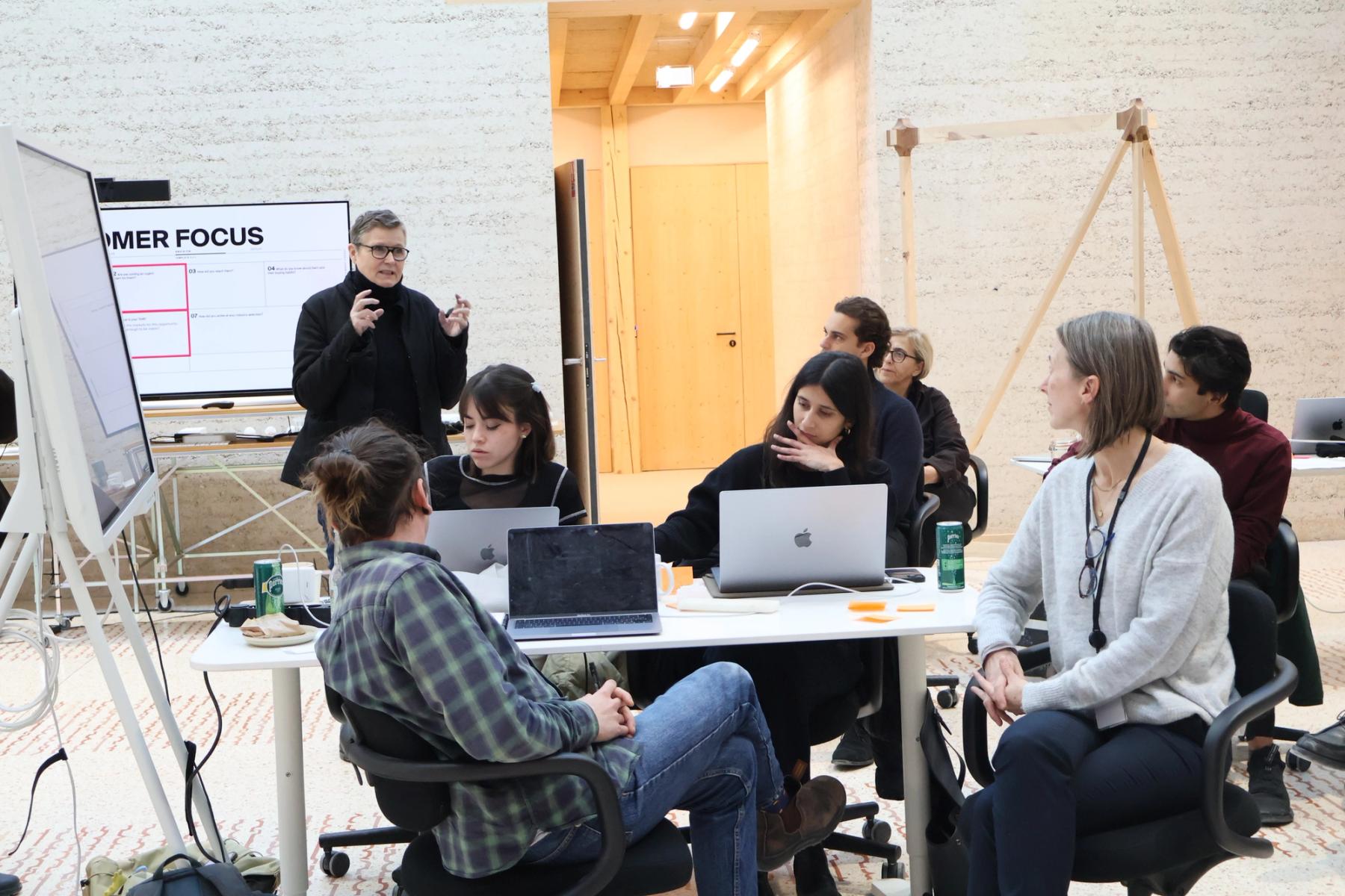 A group participates in a collaborative workshop, gathered around a table with laptops while a facilitator speaks beside presentation screens.