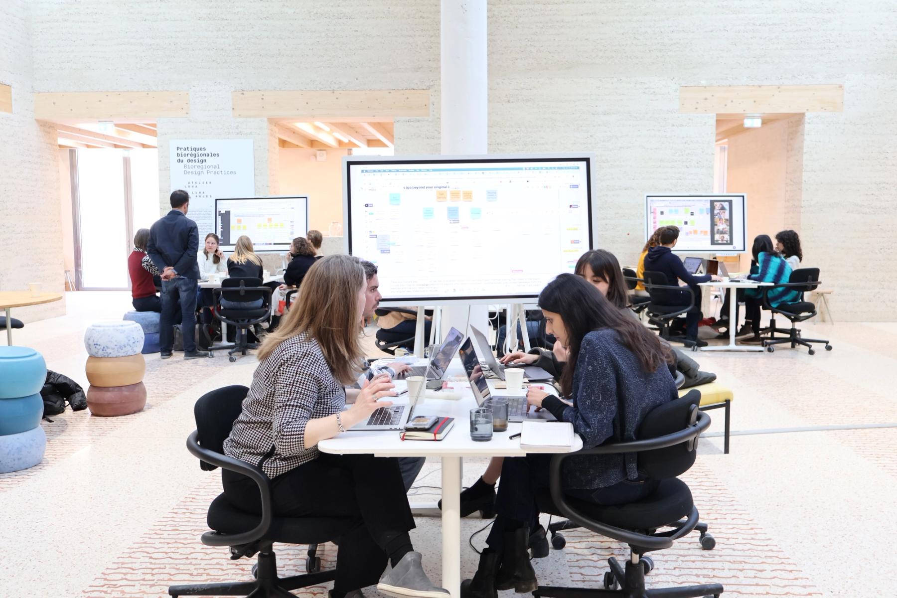 Participants collaborate in small groups around tables with laptops and digital whiteboards during a design workshop.