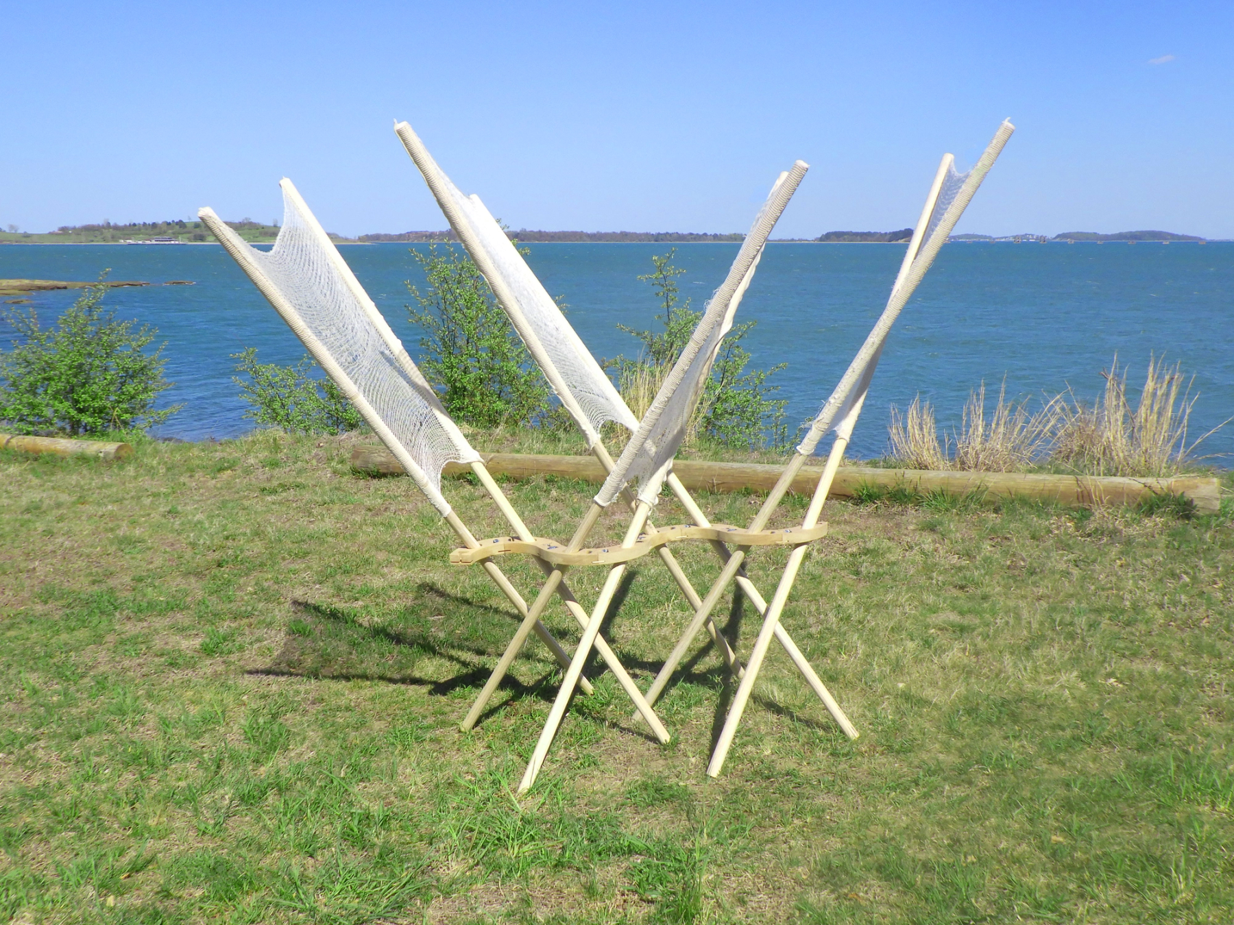 An experimental wooden structure with intersecting poles and stretched mesh panels stands on grass overlooking the water.