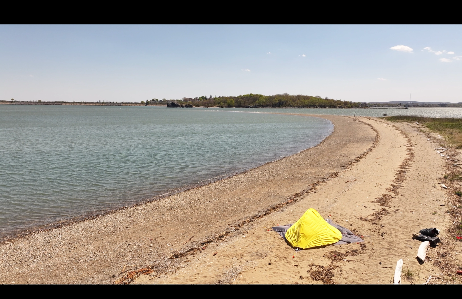 A small bright yellow experimental structure sits on a sandy shoreline beside calm water, with a curving beach and distant tree-covered land.