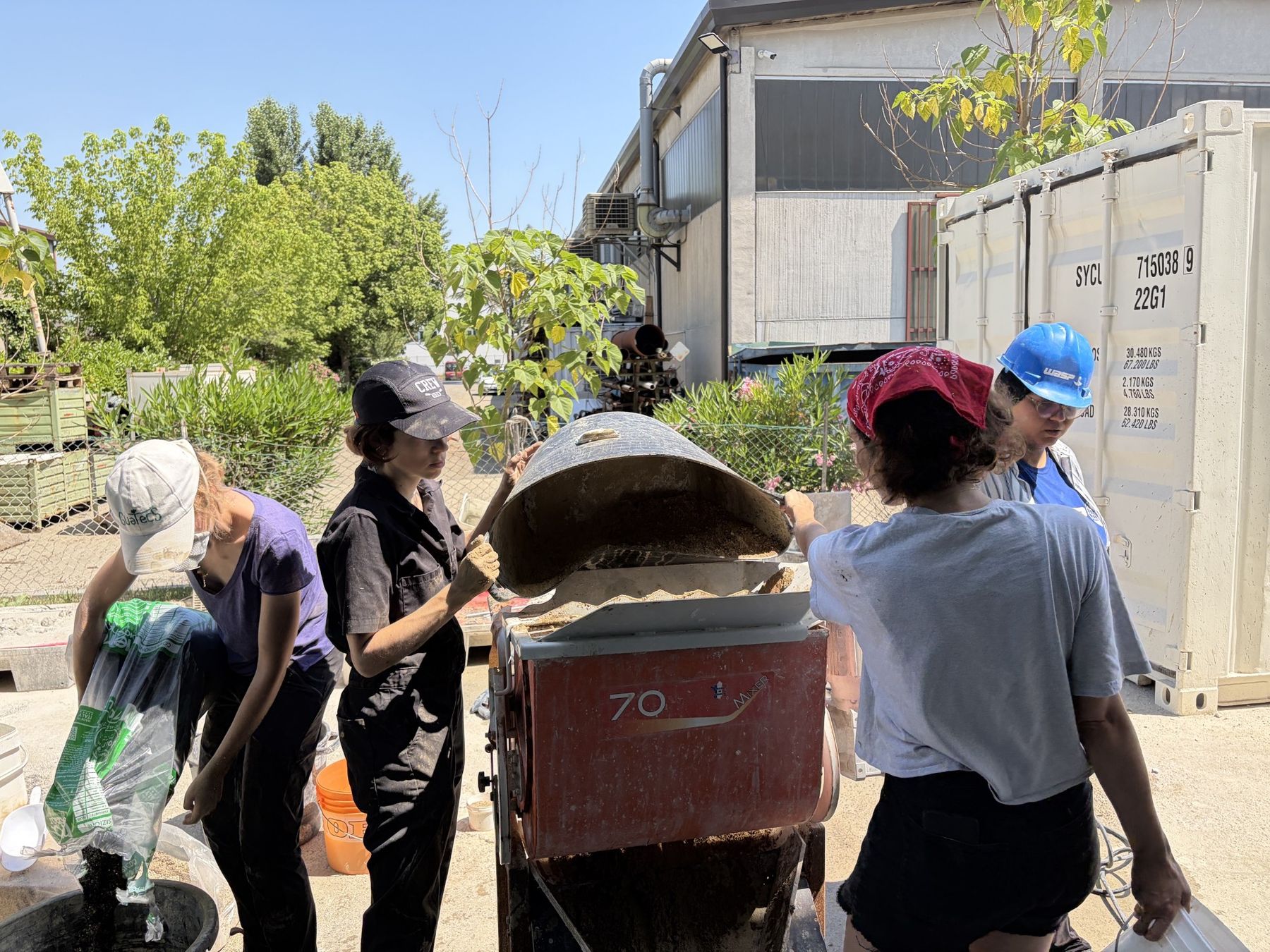 Four people work outdoors around a large industrial mixer, lifting its lid and pouring material into a container, with a shipping container and workshop building in the background.