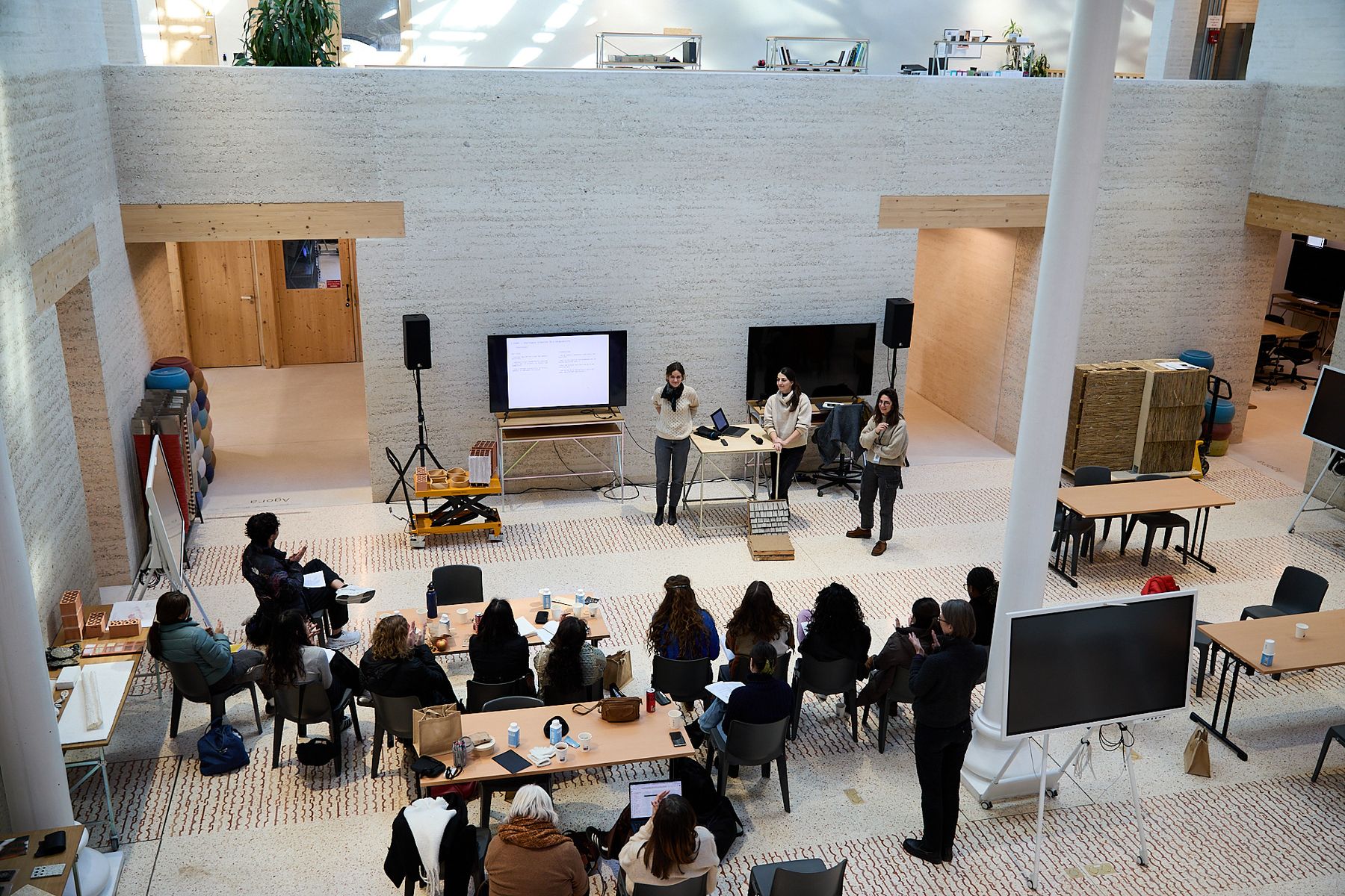 A group gathers for a presentation and discussion in a large, open workshop space, with three speakers standing at the front beside screens and material samples.