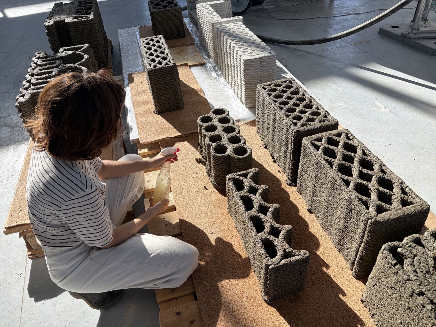 A person sprays a series of 3D-printed architectural blocks with textured surfaces and varied openings arranged on a workshop floor.