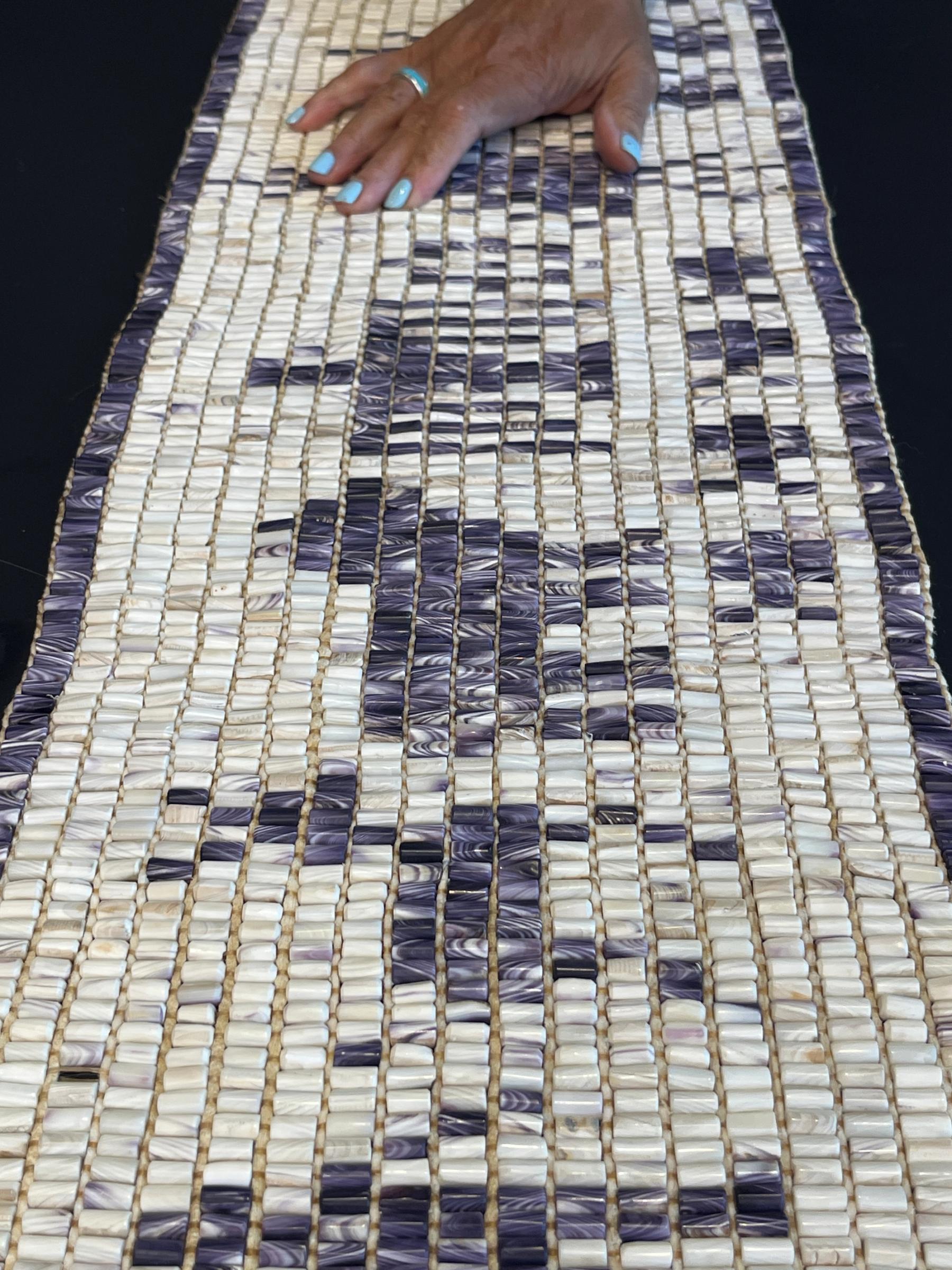 A hand rests on a woven textile composed of rows of small white and purple rectangular pieces arranged in an abstract pattern.