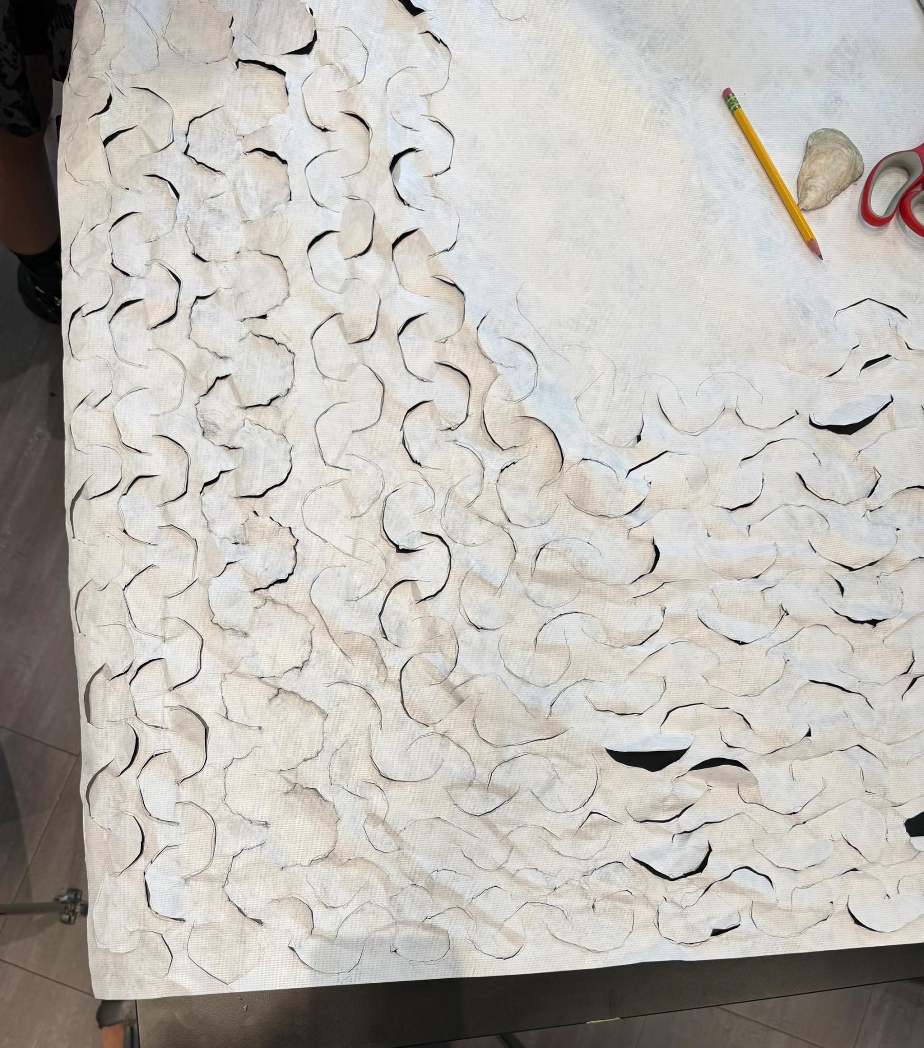Close-up of white textile material cut into repeating curved, scale-like shapes, with a pencil and scissors on the work surface.