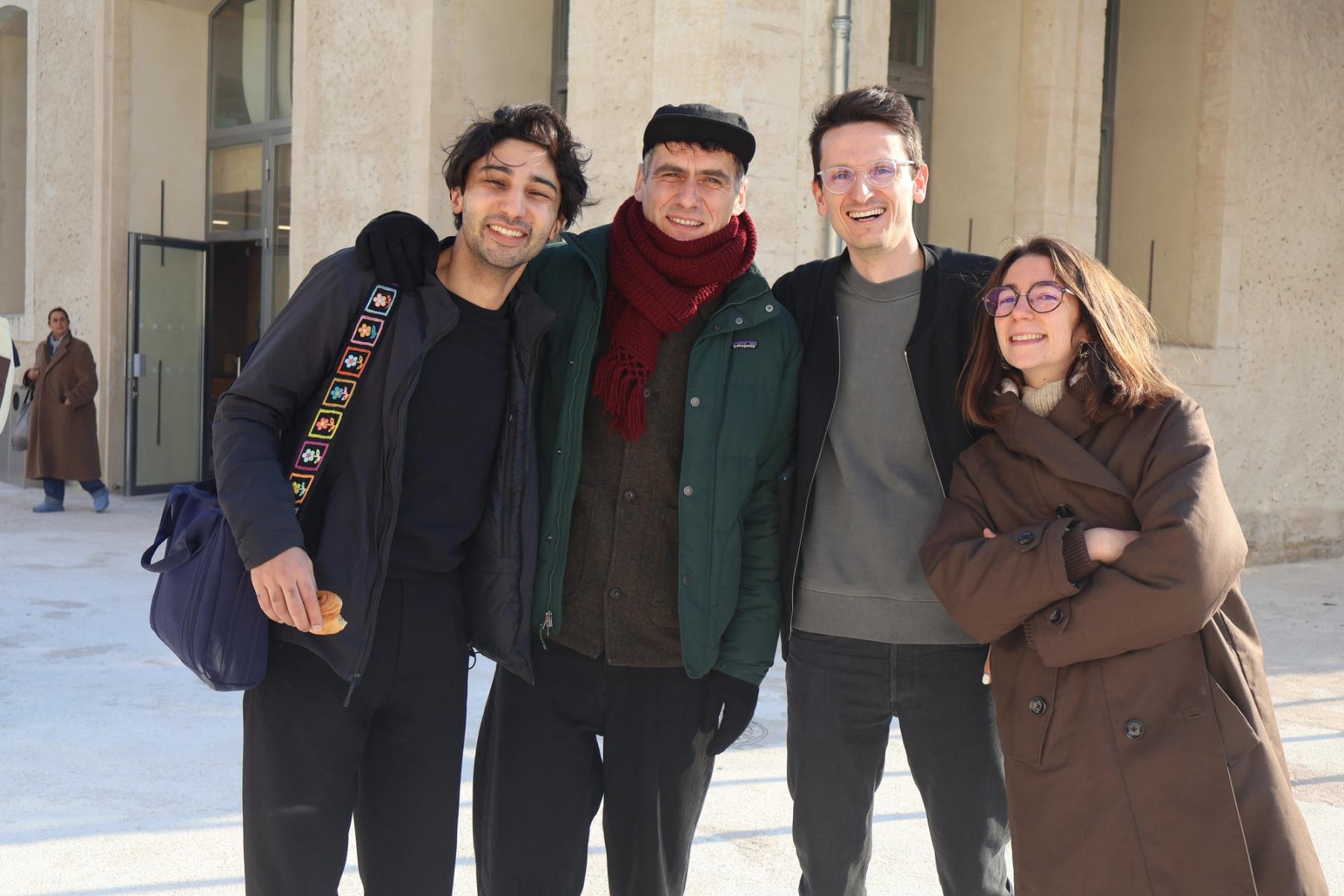 Four people pose together outdoors, smiling in front of a light stone building.