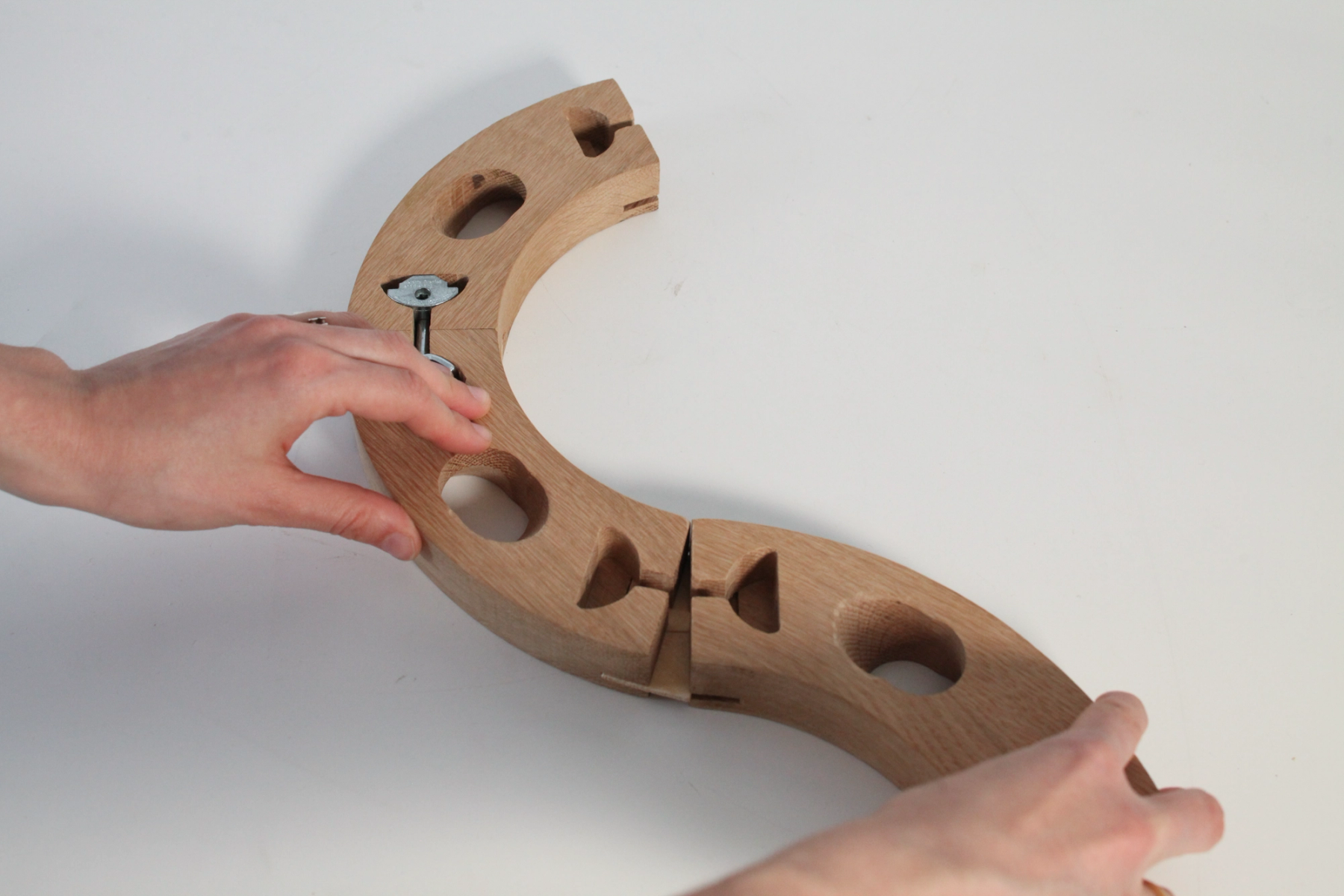 Hands manipulate a curved, modular wooden structure with circular openings and interlocking joints on a white surface.