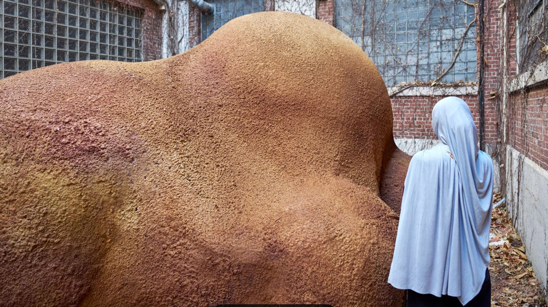 A person stands beside a large, undulating earth-toned structure with a rough, textured surface in an industrial courtyard.