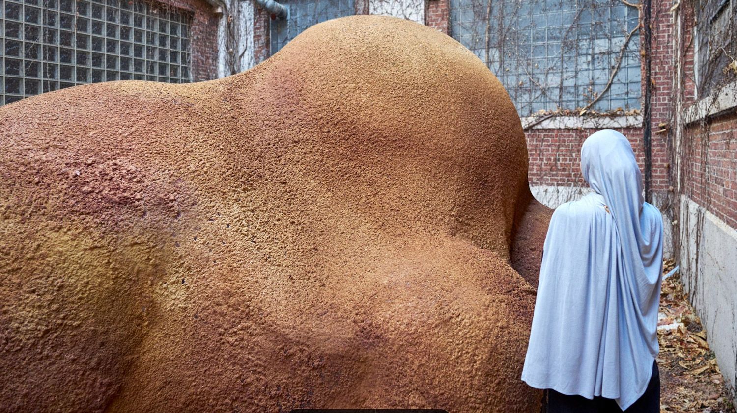 A person stands beside a large, undulating earth-toned structure with a rough, textured surface in an industrial courtyard.